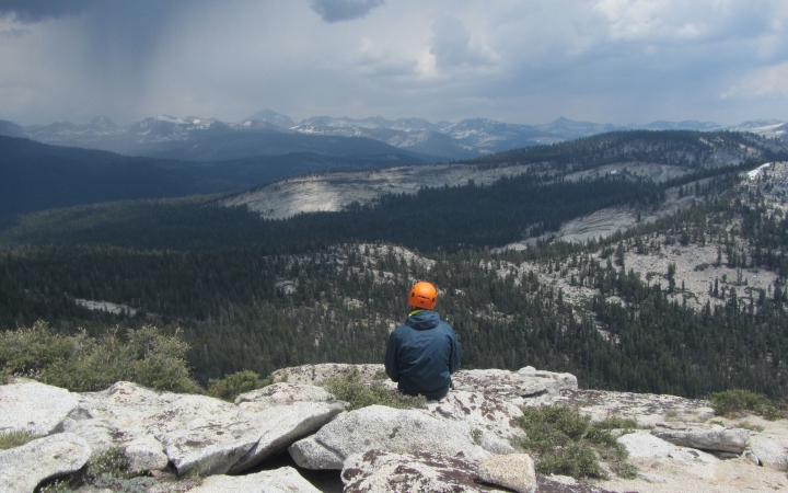 a person wearing a helmet sits on a rock high above a mountainous landscape